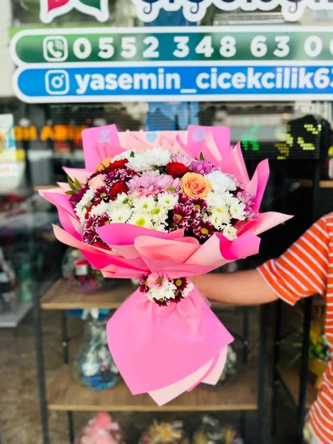 Beyaz, pembe ve bordo çiçekler arasında turuncu gül; mağaza tabelası önünde tutulan pembe ambalajlı buket.